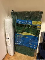 Two unopened air cleaning filter packages leaning against a wall. One is green, cut-to-fit size 20x25x1 inches, the other is a white rolled filter material.
