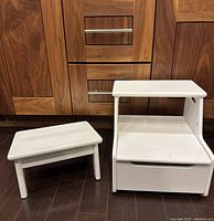 Image showing two white step stools, one smaller stool with four legs and one larger two-step stool with a drawer base, placed on dark wood flooring in front of wooden cabinets.