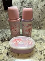 Two thermos bottles and a small lunch box container on a granite countertop, all decorated with Sanrio Bonbonribbon character and pastel colors.