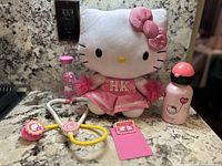 Image showing white Hello Kitty plush in pink cheerleader dress with pom-poms, toy stethoscope, small pink card and water bottle on granite surface.