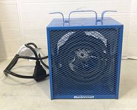 Blue metal cube shaped heater showing front protective grille and carry handle with cord laid beside it.