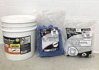 Photo of a white bucket labeled 'Nordic Xpert 202' along with two sealed plastic bags containing blue tile leveling wedges and white 2mm tile leveling spacers.