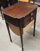 View of the sewing table showing wooden top with scratches, scalloped edges, and tapered legs with carved detail.