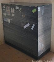 Black metal 3 drawer side filing cabinet wrapped in plastic wrap showing stickers and labels on the front and side surfaces.