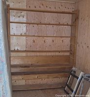 Front view of tall unfinished wooden shelf unit with five shelves against a plywood wall.