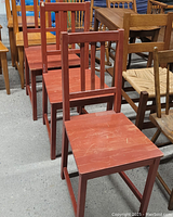 Photo showing three red stained, straight-backed wooden chairs arranged in a line with other wooden furniture in background.