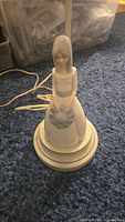 Porcelain figurine lamp showing full front view of girl holding hat, on blue carpet background.