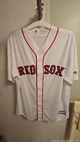 White Boston Red Sox home jersey hanging on hanger, showing front side with 'RED SOX' lettering and button-down design.