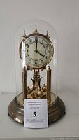 Full view of the antique German brass anniversary clock under a glass dome showing the clock face, pendulum, and brass base.