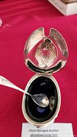 Opened Birks sterling silver mustard pot with hinged shell-motif lid, black glass liner, and matching spoon on red cloth