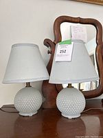 Pair of white glass hobnail pattern table lamps with light gray fabric shades, shown on a wooden surface in front of a mirror. One shade is distorted and bent.