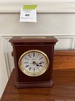 Front view of the wooden mantel clock showing the clock face, gold-tone bezel, and removable name plate on top.