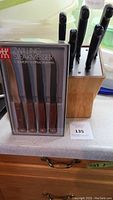 Wooden knife block with black-handled knives and one boxed set of Zwilling steak knives with wooden handles shown on a countertop.