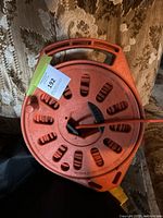Electric cord wound on orange plastic spool with handle, yellow plug end visible, spool lying on floral fabric background.