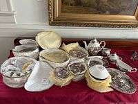 Full lot displayed on red tablecloth with framed wall art background. Multiple dishes arranged showing variety of plates, bowls, teapot, gravy boat, sugar and creamer.