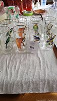 Overview of eight clear glass tumblers with gold rims and colorful bird illustrations