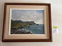 Framed painting showing a coastal scene with rocky shore and cliffs with waves hitting the shore under cloudy sky.
