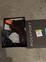Photo showing one chocolate brown Rockport men's boot inside a black Rockport box with lid displaying brand name.