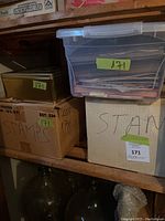 Four storage containers on wooden shelf: clear plastic bin, two cardboard boxes, metal tin