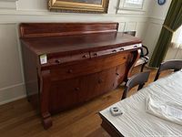Front view of a vintage American Empire style mahogany credenza buffet placed in a dining room showing four small drawers on top and two large cabinet doors below, polished wood surface and curved scrolled legs.