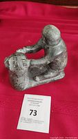 Front angled view of the soapstone sculpture of an Inuit figure gripping a seal on a red cloth background with auction lot number card visible.