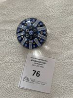 Top-down view of round blue Strathearn art glass paperweight with Millefiori and Maltese cross design, displayed on a light textured surface next to an auction lot number card.