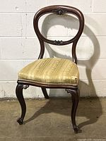Front angled view of antique balloon back chair showing carved wooden frame and beige striped upholstered seat with wear on corner.