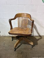 Front view of antique oak office chair showing curved backrest with slats, wooden arms, flat wooden seat, and caster wheels on oak five-legged base.