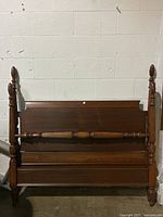 Front view of the antique walnut four post double bed frame mounted against a white brick wall showing the headboard, footboard, and posts with turned details.