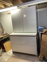 Front view of the white storage pantry cupboard showing two upper cabinet doors and two lower drawers in a basement or garage setting.