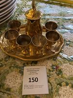 Photo showing brass coffee pot, 6 cups and tray from above and front angle on a floral tablecloth. Auction lot card number 150 included.