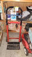 Full view of red and black metal dolly hand truck standing upright in a workshop area, surrounded by various items and shelving.