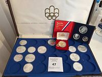 Open presentation case showing white satin lid with gold Olympic rings logo, blue velvet coin tray with silver commemorative coins and red presentation box