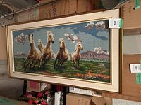 Framed needlepoint artwork showing five running horses with a natural background, under glass cover and light wood frame.