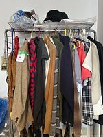 Rack of men's semi formal and casual wear clothes, including blazers, jackets, sweaters, shirts, and accessories in bags on top of rack.