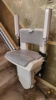 Close-up of stairlift seat with gray padded seat and backrest, white armrests, and base unit attached to the rail.
