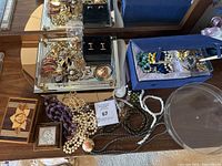 Wide view of assorted costume jewelry on mirror tray and nearby surface showing necklaces, brooches, earrings, and boxed cufflinks.
