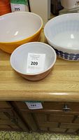 Three vintage Pyrex mixing bowls on a wooden surface. Small brown bowl with white wheat pattern, medium yellow bowl, and larger white bowl with blue dots.