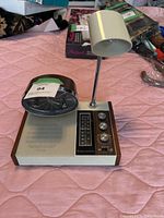 Photo of the Longines Symphonette AM/FM radio with attached alarm clock and lamp, showing the front and side view on a pink quilted surface.