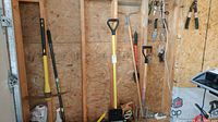 Photo showing full-sized long-handled garden tools including shovel, pickaxe, pitchfork, and others mounted against a wall.