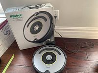 iRobot Roomba vacuum robot alongside its original box on wooden floor, box shows product image and branding.