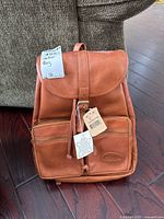 Front view of tan Bugatti genuine leather knapsack backpack showing flap cover, buckle closure, drawstring, and two front zippered pockets with tags intact.