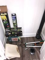 Photo showing five boxed solar stake lights leaning against wall, garden loppers and pruning tools laid on floor with spool of wire and cardboard box
