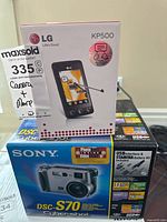 Photo shows Sony DSC-S70 Cyber-shot camera box with product image and feature callouts, alongside LG KP500 touchscreen phone box.