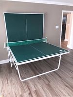 Overall image showing table tennis table half folded upright against wall, wooden floor, beige walls.