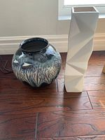 Two ceramic vases on hardwood floor by window: one white tall geometric faceted, one round blue cream black glazed with nature pattern