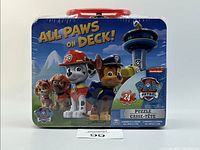 Front view of the tin lunch box packaging showing Paw Patrol character illustrations and puzzle details.