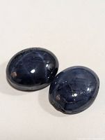 Two polished oval cabochon blue star sapphire gemstones on white background, showing asterism and internal features.