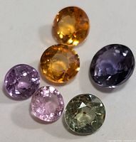Close-up photo of six faceted sapphires in various colors including yellow-orange, purple, pink, and green, arranged on a white surface.