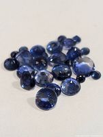 Close-up photo showing multiple faceted blue sapphire gemstones in various sizes and cuts on a white background.
